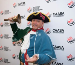 Man in colonial dress ringing a bell in front of a wall of CAASA logos